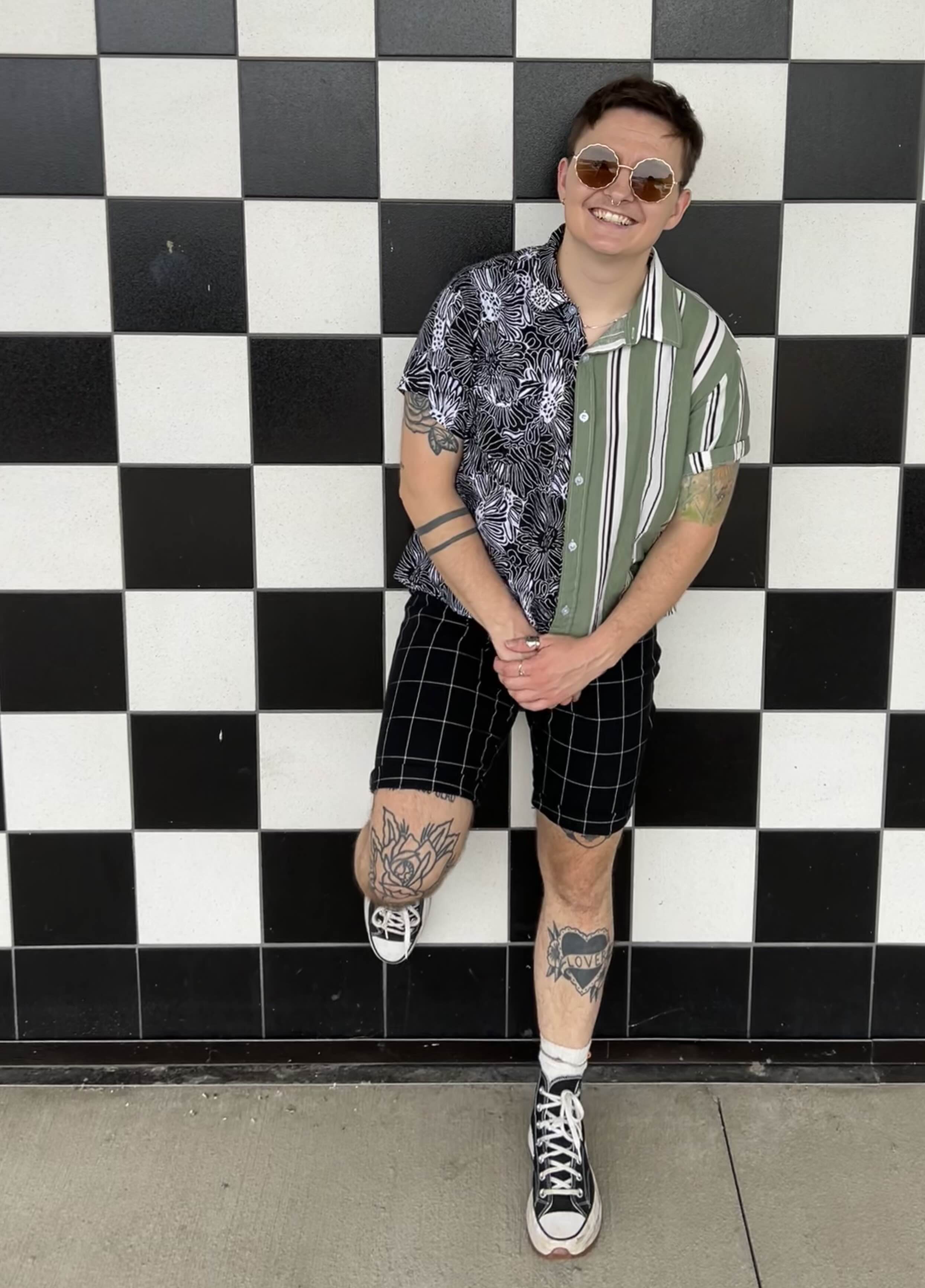A smiling Michelle stands with one foot on the wall behind them and their hands clasped in front of them. Michelle is white with tattoos and short brown hair. They are wearing sun glasses, plaid shorts, and a button up short sleeve shirt with two prints, a green and white stripe and a black and white paisley print. The background is black and white checkered.